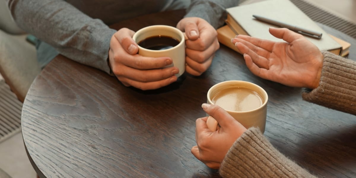 Two people talking over cups of coffee at a table, one gesturing as they explain something. Two people talking over cups of coffee at a table, one gesturing as they explain something.