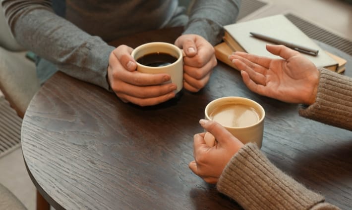 Two people talking over cups of coffee at a table, one gesturing as they explain something.
